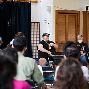 Jules, a fat white queer wearing all black, sits in a room with others, one hand on their chest and another on their belly teaching “Singing with Our Whole Selves” at Let My People Sing, Summer 2023